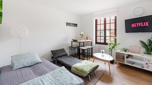 Une chambre moderne et bien éclairée, avec deux lits, une petite table agrémentée d’une plante, un téléviseur et une fenêtre équipée de stores.