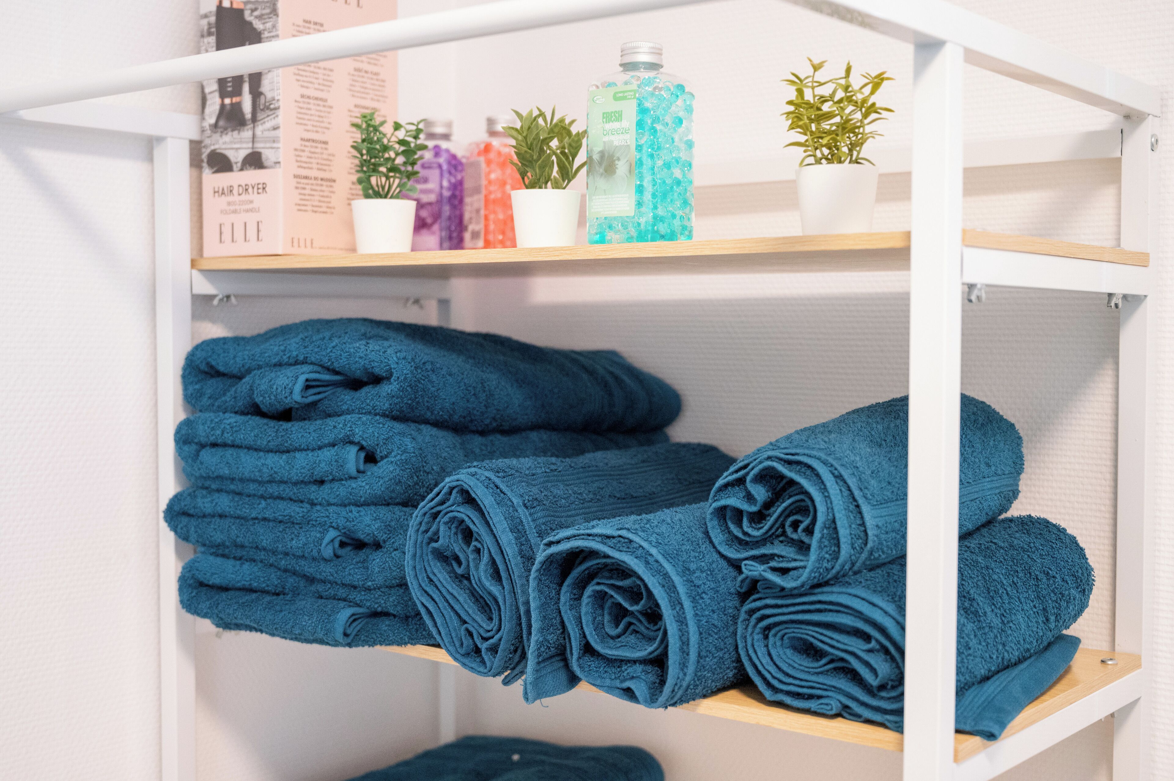 A shelf with folded blue towels, potted plants, and bottles of hair spray.