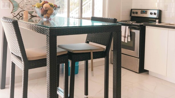 A modern kitchen with white cabinets, a stainless steel stove, and a microwave. There is a dining table with wicker chairs and a bowl of fruit. - Venetian Breeze Cozy Studio #7 (Venetian Road Settlement)