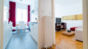 Una cocina moderna con armarios blancos, una zona de comedor con una mesa y sillas blancas, una sala de estar con un sofá blanco y un televisor sobre una repisa de madera.