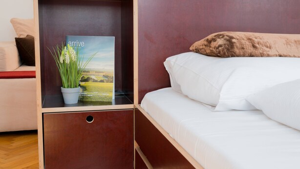 Una cama bien hecha con ropa blanca y una almohada marrón, una mesita de noche pequeña con una planta en maceta y un libro titulado "arrive", y una mesita de noche de madera con un cajón.