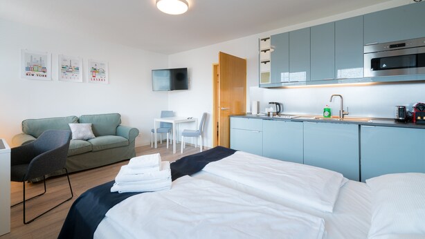 Ein modernes Hotelzimmer mit Bett, Schreibtisch, Sessel, einer kleinen Küchenzeile und einem Flachbildfernseher.