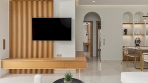 A modern living room with a flat-screen TV mounted on a wooden panel wall, a round coffee table with candles and a potted plant, and a dining area with a round table and chairs. - Adore Gold Natural Retreat by Estia (Perama)