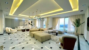 A spacious living room with a large sectional sofa, a coffee table, and a TV mounted on the wall. The room has a high ceiling with recessed lighting and a view of the outdoors through large windows. - Nile view appt, Cornish al Nile (Cairo)
