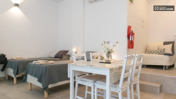 Una habitación pequeña y limpia con dos camas, una mesa con sillas, un jarrón con flores y un extintor en la pared.