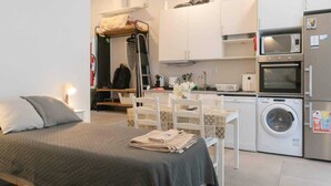 Un dormitorio compacto con cama, mesita de noche y una pequeña kitchenette equipada con microondas, horno y lavadora.