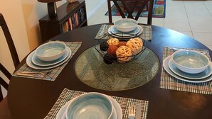 Un comedor con una mesa redonda dispuesta para cuatro personas, rodeada de sillas. Se ve un sofá y un televisor al fondo.