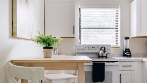 Uma cozinha com armários brancos, fogão, chaleira, liquidificador, ventilador de teto, janela com persianas, uma cadeira e uma planta.