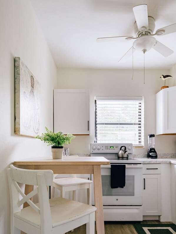 A kitchen with white cabinets, a stove, a kettle, a blender, a ceiling fan, a window with blinds, a chair, and a plant.