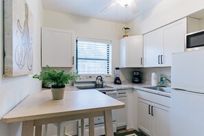A compact kitchen with white cabinets, a small dining table, a kettle, a coffee maker, and a potted plant.