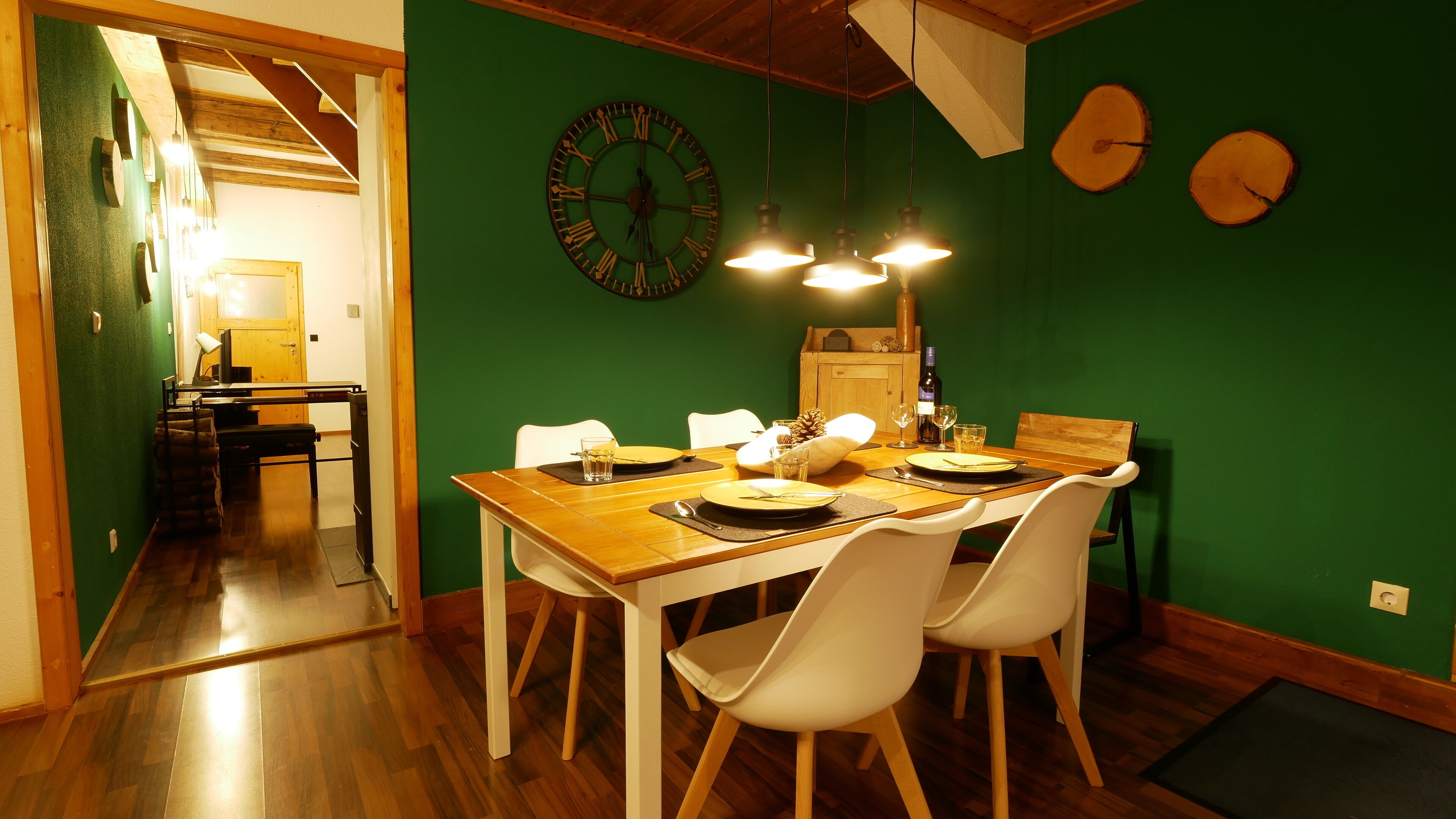 A dining area with a wooden table set for two, white chairs, and a green wall with a clock.