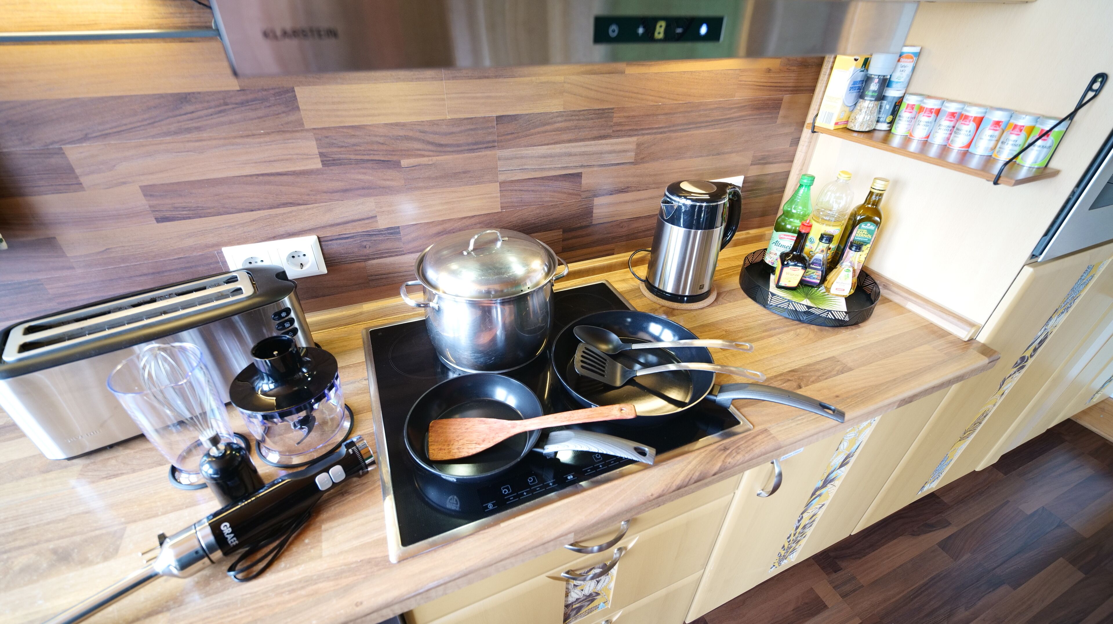 Una cocina con encimera de madera, estufa, cafetera, olla, sartén, cuchara de madera y especiero.