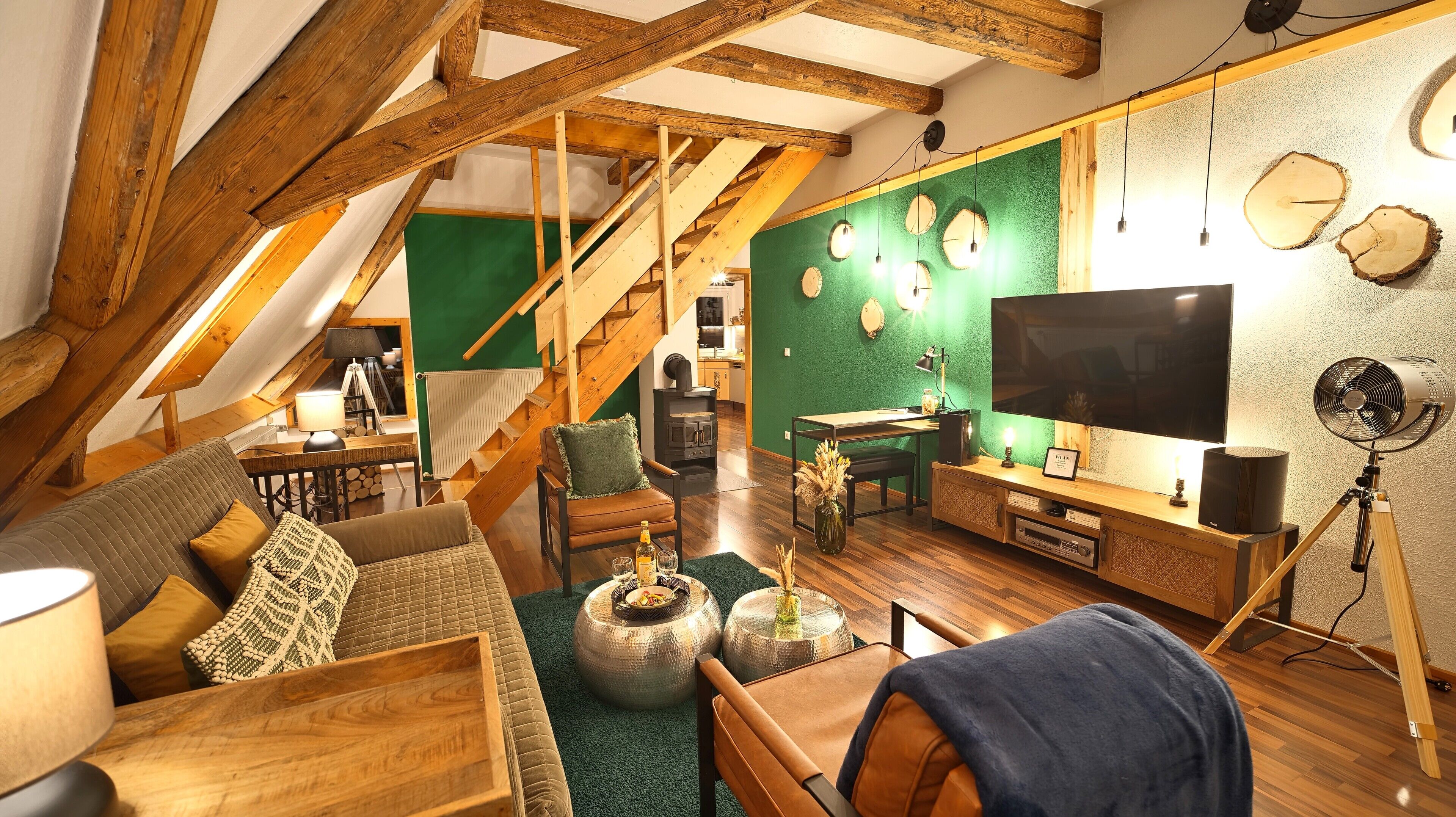 Un salon chaleureux de style loft, avec des poutres apparentes, un mur d’accent vert, un grand téléviseur à écran plat et un meuble de rangement en bois.