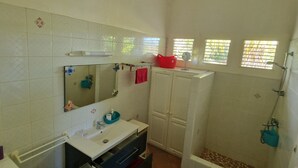 A bathroom with a white sink, mirror, and tiled walls. There is a shower area with a window and a red bath mat. - Studio Alamanda Terrasse Face à la Piscine (sainte rose)