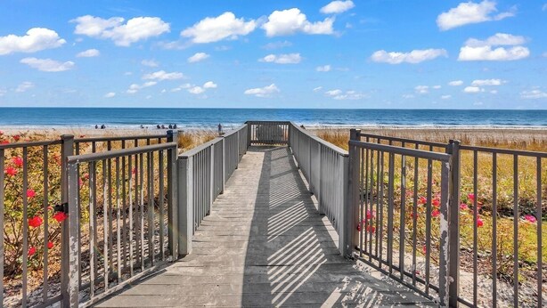 Una passerella di legno che conduce a una spiaggia, con cielo sereno e vista sull'oceano in lontananza.