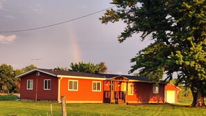 Une maison rouge avec un porche à l’avant et un garage, entourée d’un champ verdoyant et d’un grand arbre.