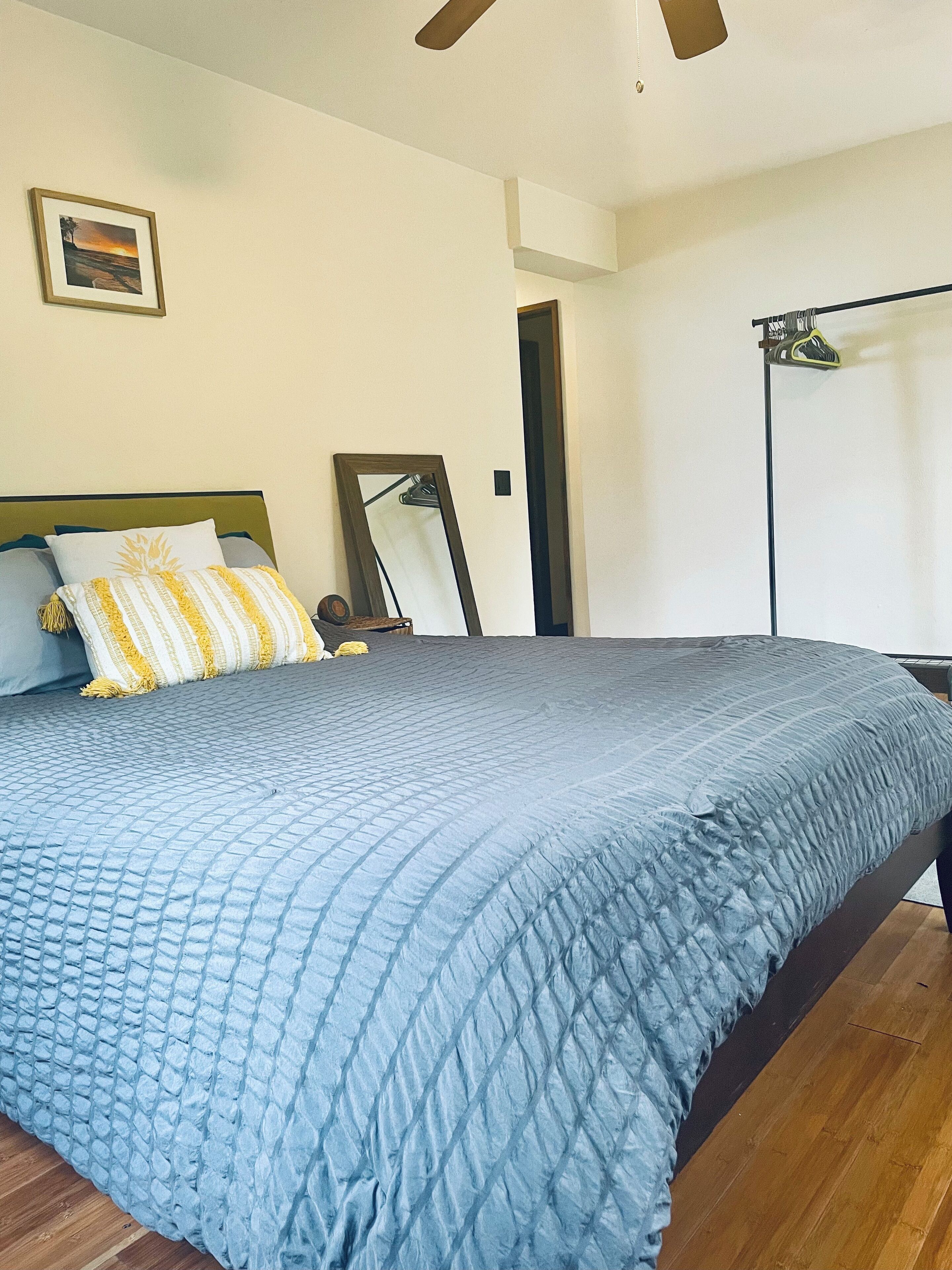 A bedroom with a bed, a ceiling fan, a framed picture on the wall, and a door leading to another room.