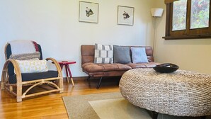 Un salon avec un plancher en bois, un canapé en cuir marron, un pouf en osier, un fauteuil en osier et une petite table rouge. Deux tableaux encadrés sont accrochés au mur.