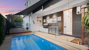 Un área de piscina exterior moderna con cerramiento de vidrio, zona de barbacoa y cocina equipada con refrigerador y armarios.