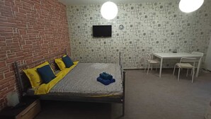 Un dormitorio con pared de ladrillo, cama con almohadas amarillas y azules, un escritorio con dos sillas y un televisor en la pared.