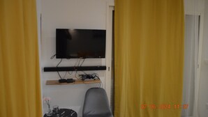 Una habitación con un televisor montado en la pared, un pequeño estante debajo y una puerta con cortinas amarillas.