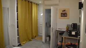 Cocina compacta con microondas, cafetera y una mesita. Una cortina amarilla cubre parcialmente una puerta que da a un baño.