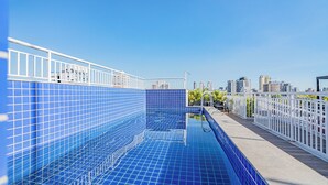 Una piscina en la azotea con baldosas azules, barandilla blanca y un paisaje urbano de fondo.