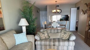 A living room with a beige sofa, a striped sofa, a round coffee table with a decorative bowl, a large plant, and a dining area in the background.