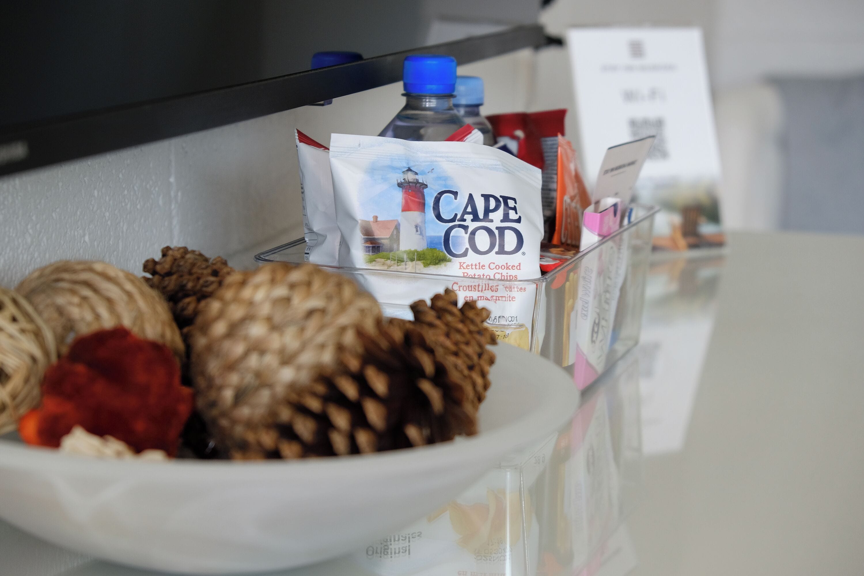A hotel room with a white table displaying a plate of nuts, a bottle of water, and a bag of Cape Cod Kettle Cooked Potato Chips.