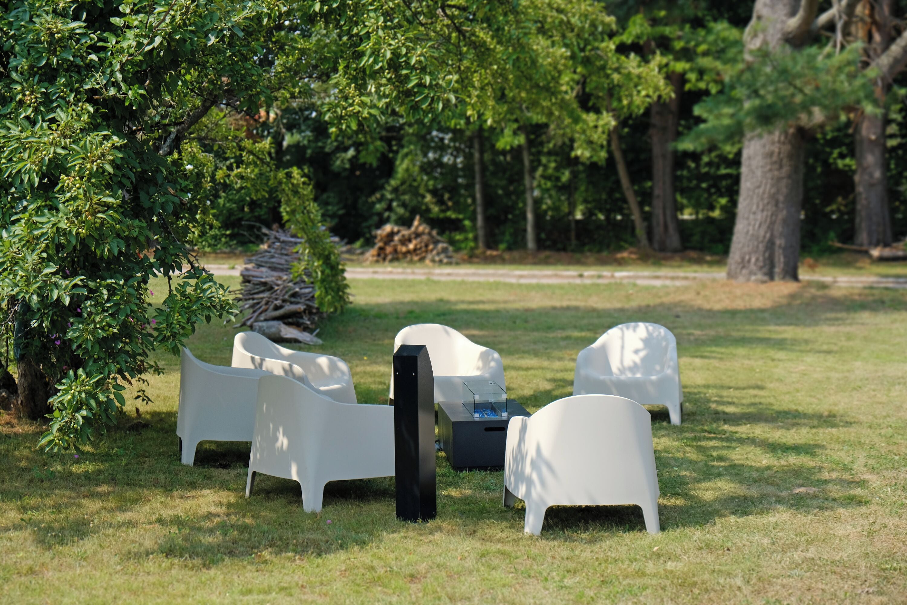 Outdoor seating with white chairs and a black table, surrounded by grass and trees.