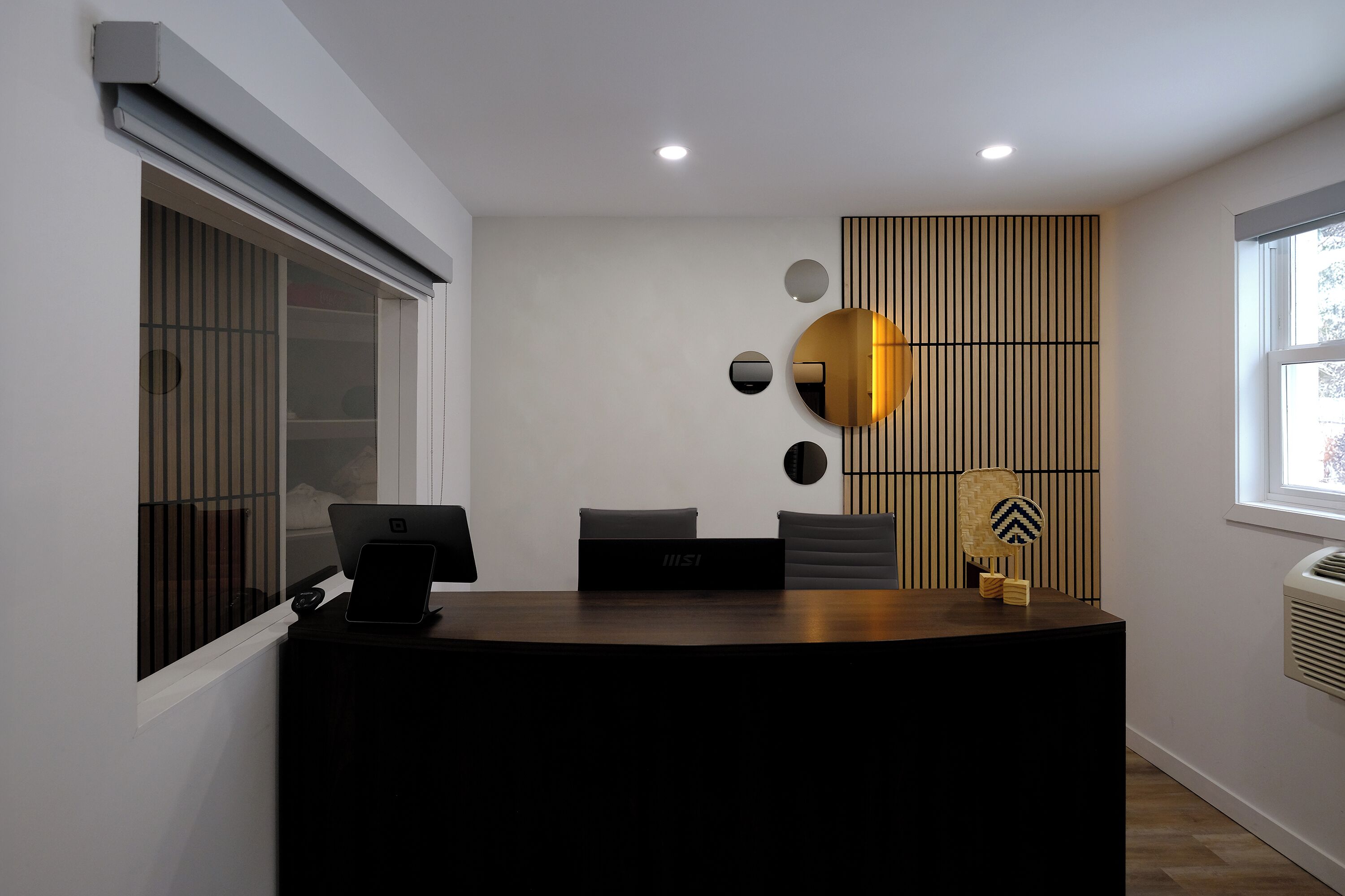 Un mostrador de recepción con una computadora portátil, un teléfono y una pequeña mesa con un objeto decorativo. Hay tres sillas y una pared con líneas verticales y espejos circulares.