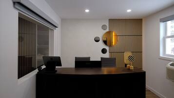 Un mostrador de recepción con una computadora portátil, un teléfono y una pequeña mesa con un objeto decorativo. Hay tres sillas y una pared con líneas verticales y espejos circulares.