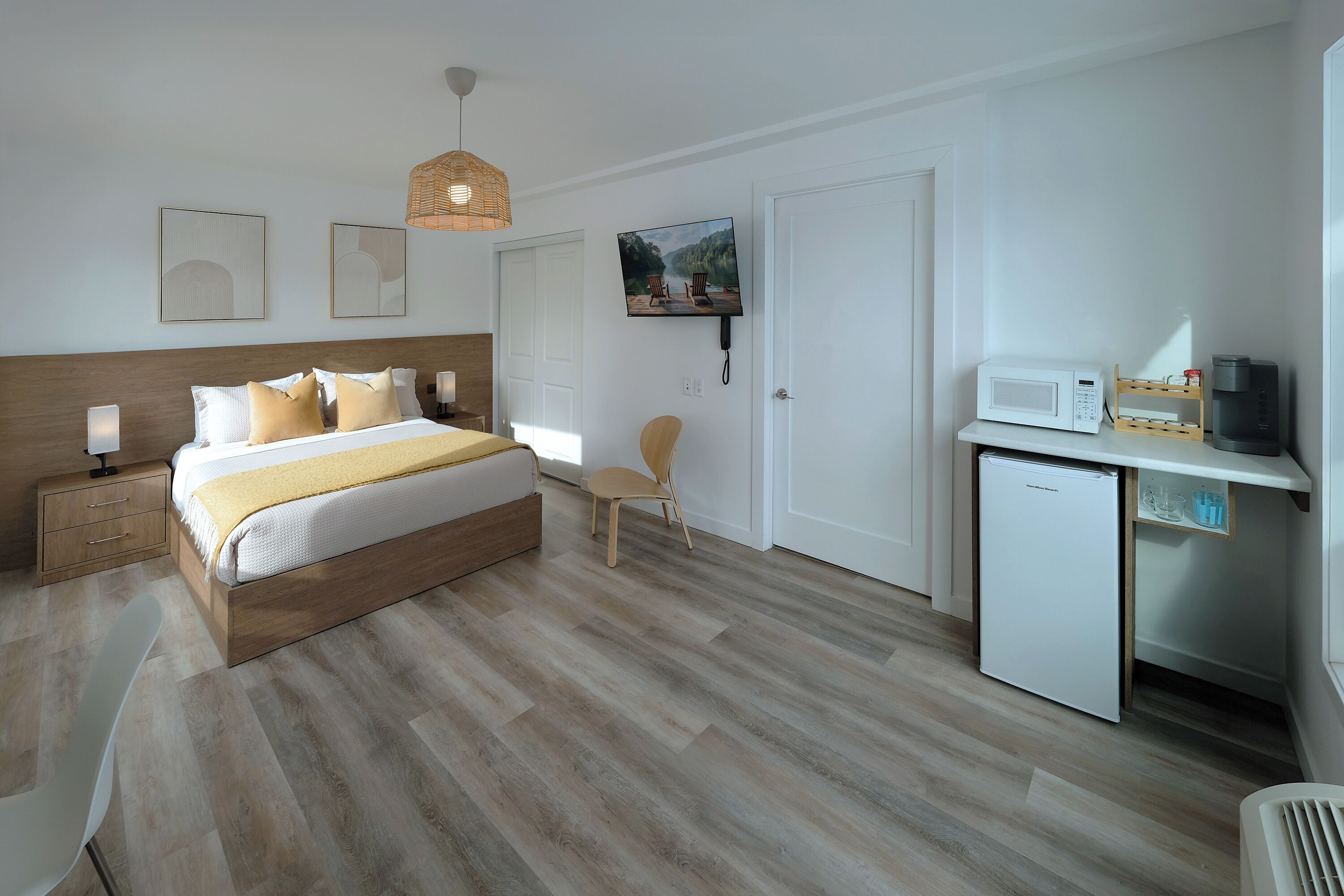 Un dormitorio moderno con cama de madera, mesitas de noche, una silla, televisor y una pequeña kitchenette con microondas y cafetera.