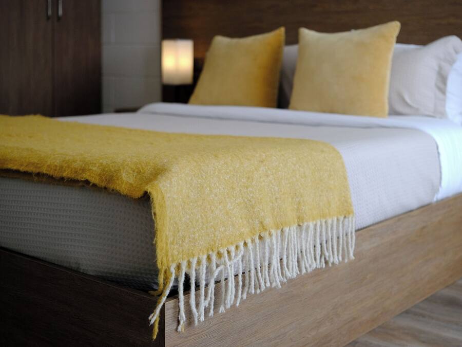 A bed with a yellow throw blanket and white pillows in a room with wooden flooring and a nightstand.