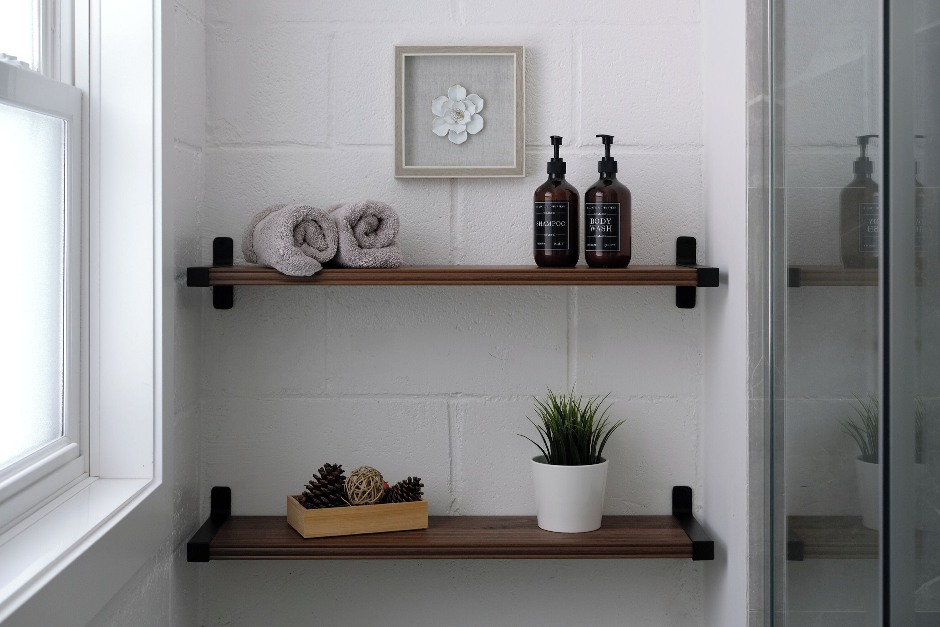 A bathroom with two wooden shelves, one holding rolled towels and the other with shampoo and body wash bottles. A framed flower decoration hangs above the shelves, and a potted plant is on the lower shelf.