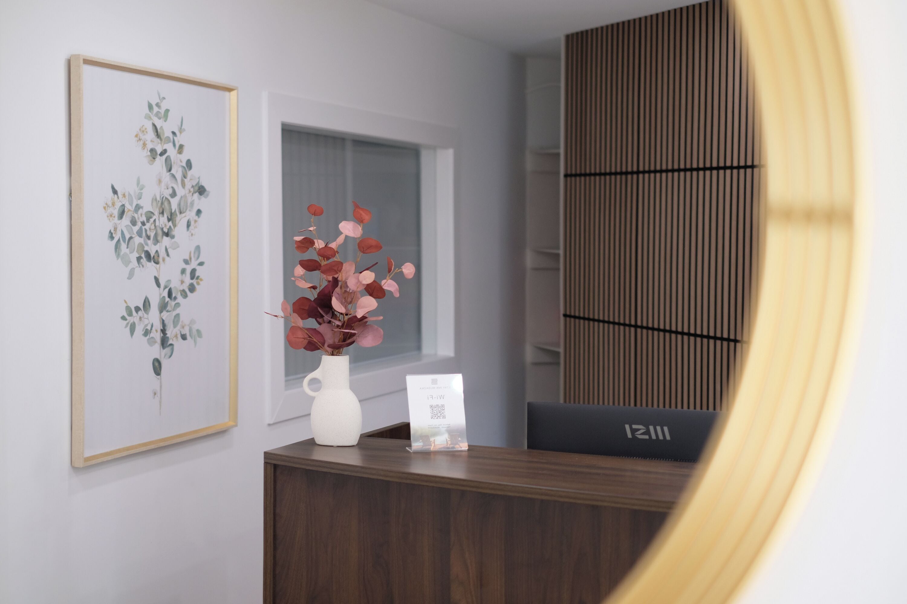 A reception desk with a framed botanical print, a vase with pink flowers, and a QR code sign.