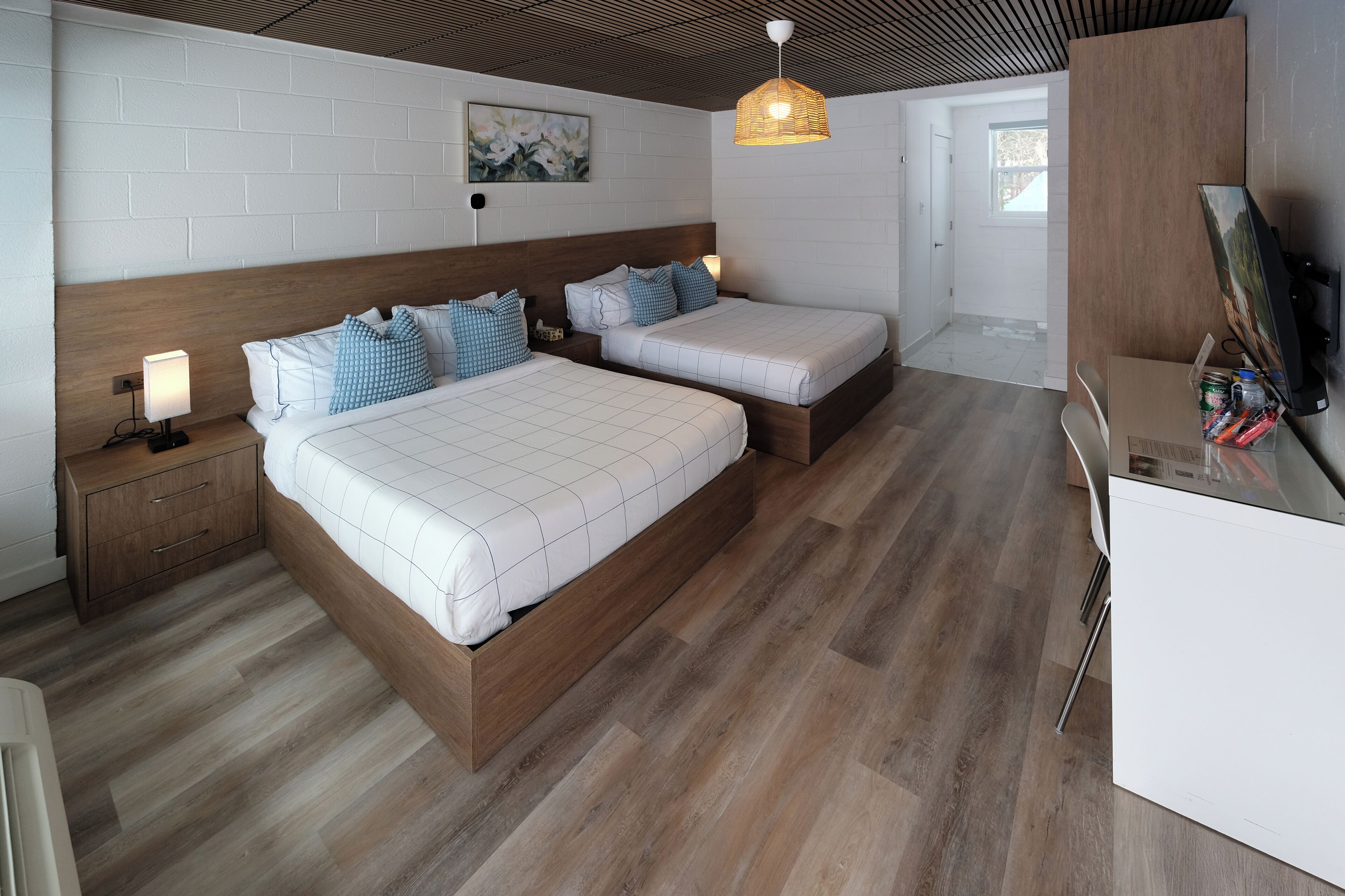 A modern bedroom with two beds, a wooden headboard, a nightstand, a desk with a computer, and a wall-mounted TV.