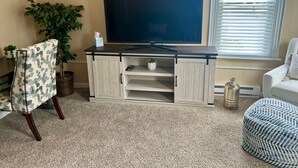 Un soggiorno con una TV a schermo piatto su un mobile in legno, una pianta in vaso, una poltrona fantasia e una finestra con persiane.