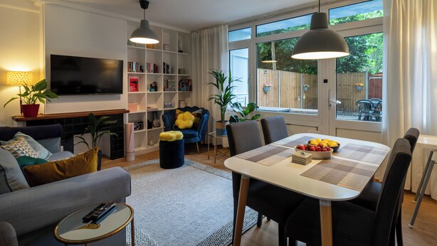 A modern living room with a TV, bookshelf, dining area, and large windows.