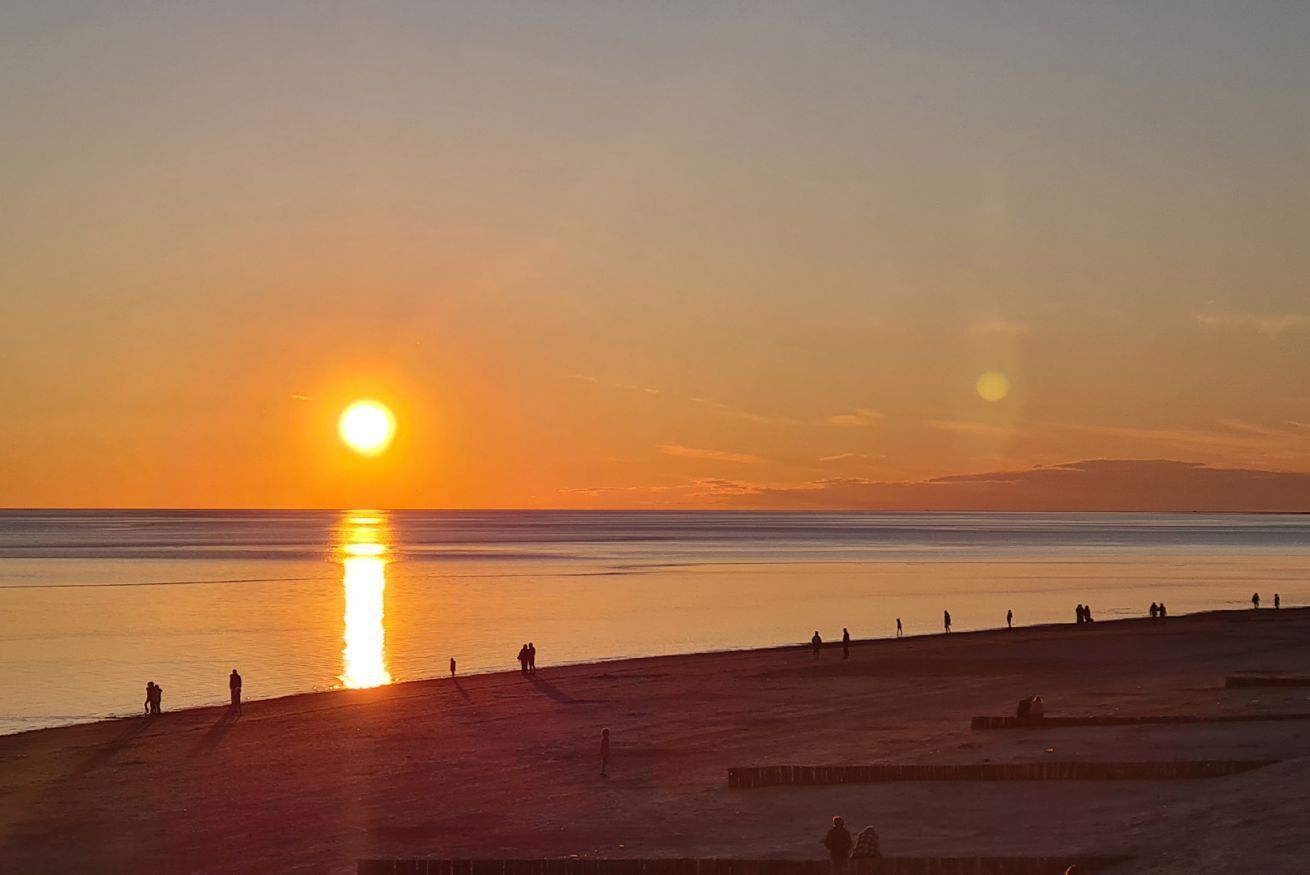 Cena de praia com o sol se pondo sobre a água, pessoas caminhando pela orla e céu claro.