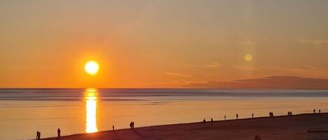 Cena de praia com o sol se pondo sobre a água, pessoas caminhando pela orla e céu claro.
