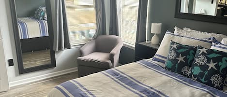 A bedroom with a large bed, a chair, a nightstand, a mirror, and a view of the outside through the window.