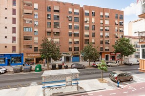 Un immeuble à plusieurs étages, doté d’une façade en briques, d’une rangée de fenêtres, et d’une rue devant, où sont garées des voitures et où se trouve un arrêt de bus.