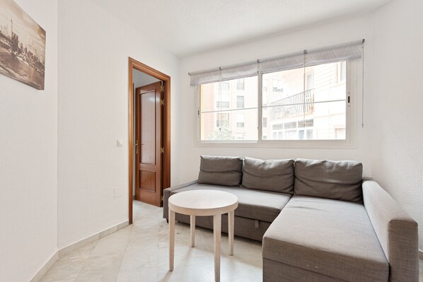 Un salon comprenant un canapé, une table ronde, une porte et une fenêtre donnant sur des immeubles.