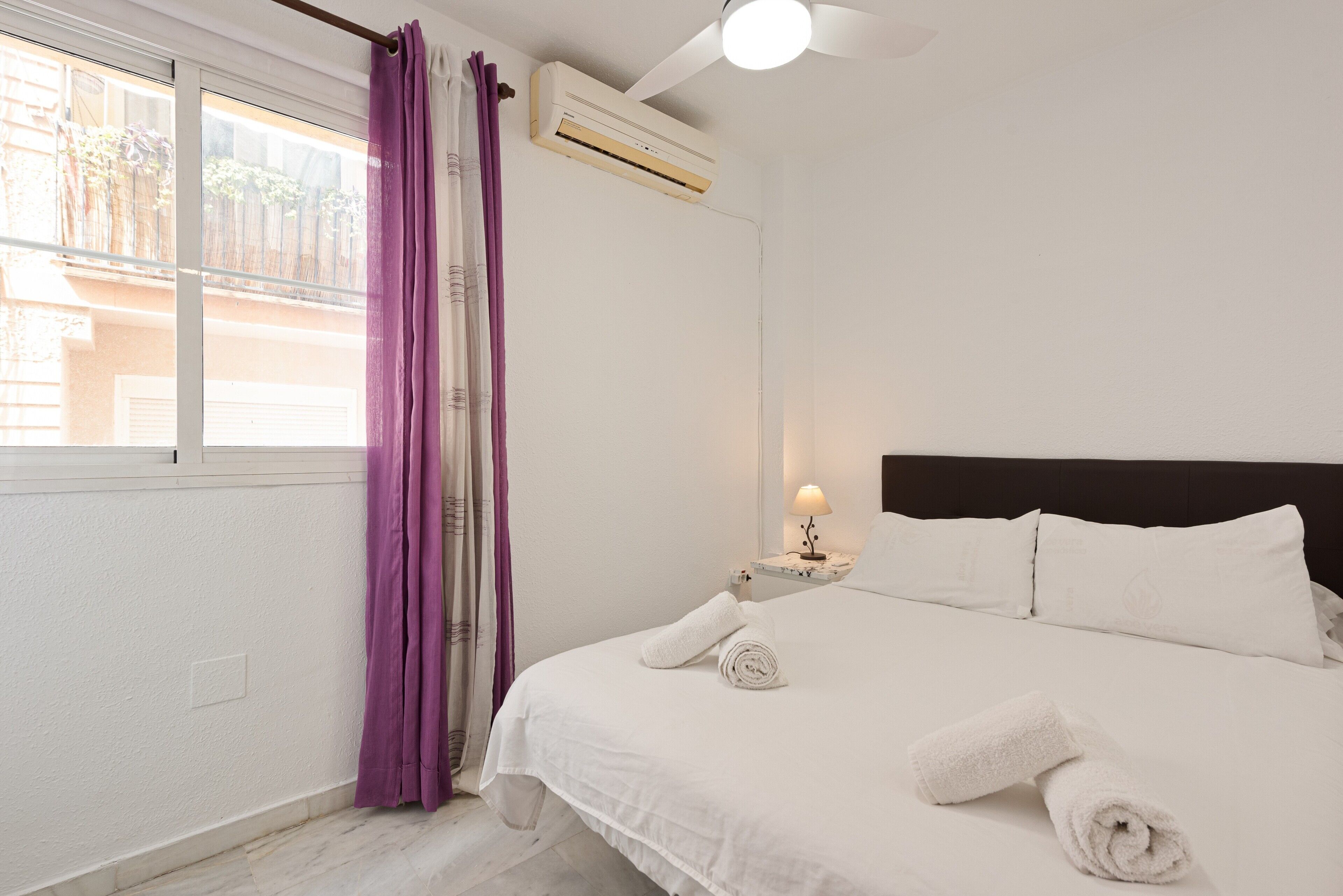 Hotelkamer met een bed, nachtkastje met lamp, airconditioning, raam met gordijnen en uitzicht op gebouwen.