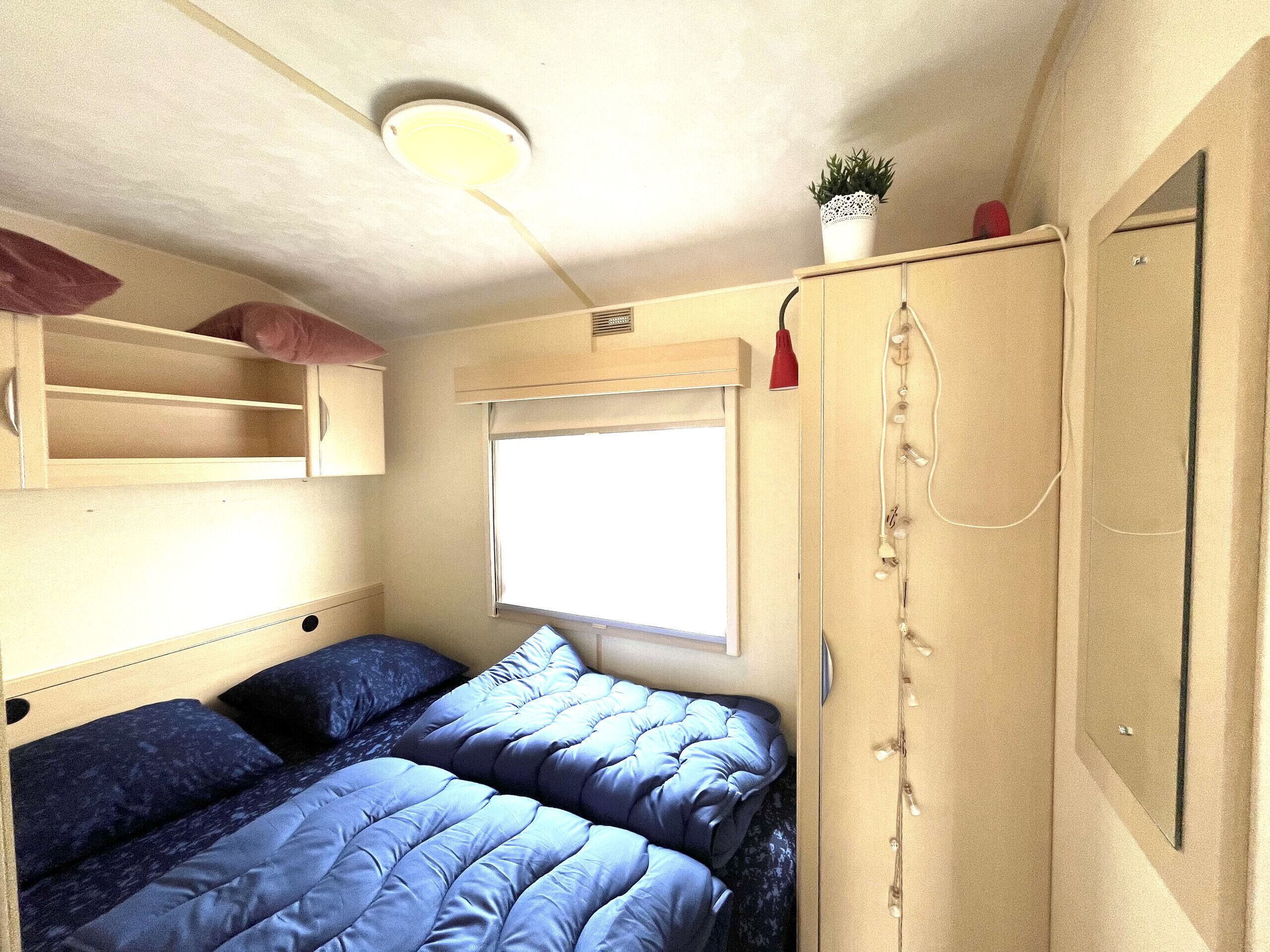 Quarto compacto com cama, janela, guarda-roupa e prateleira com uma planta.