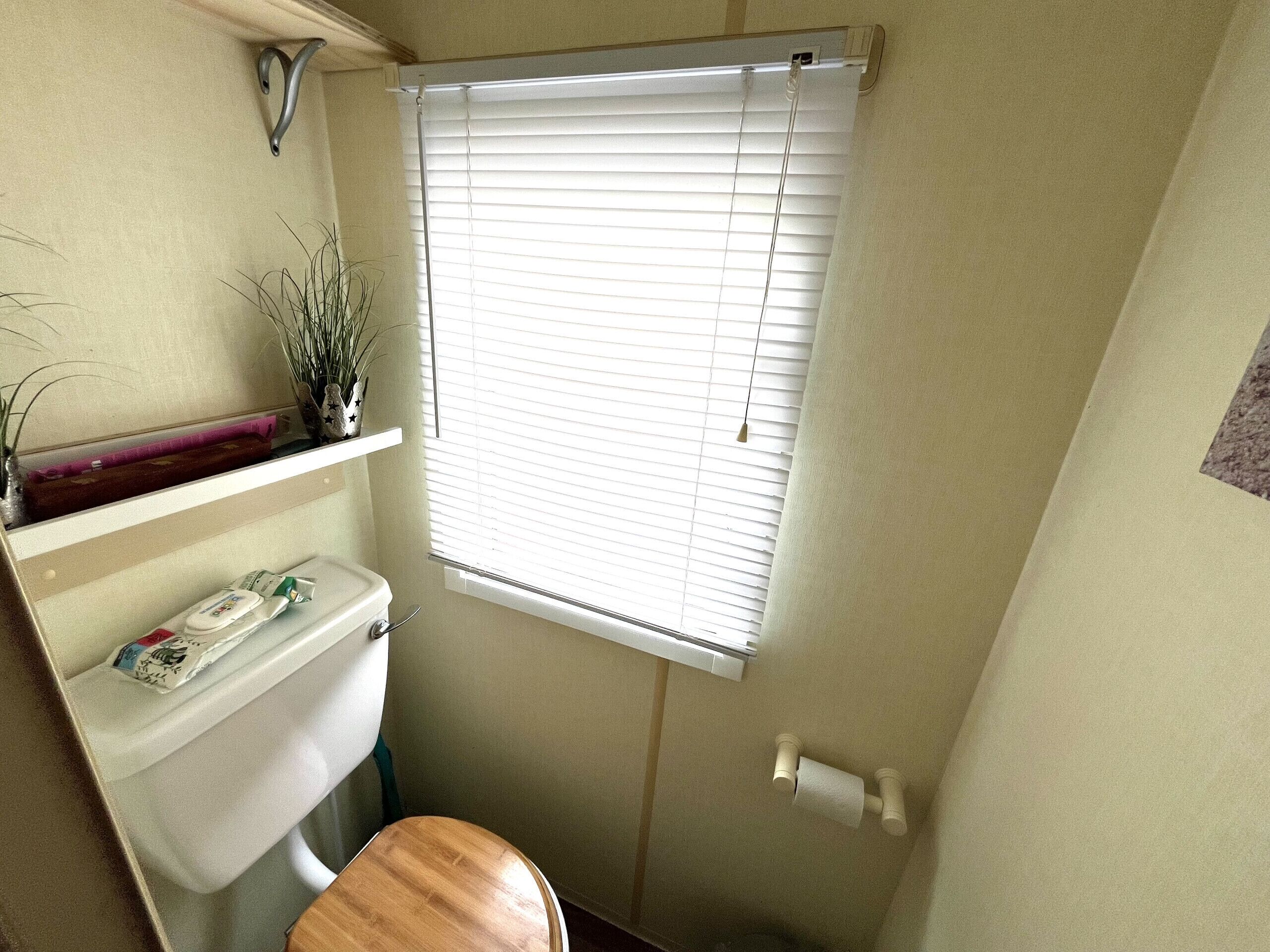 A small bathroom with a toilet, a wooden seat, a shelf with toiletries, and a window with blinds.