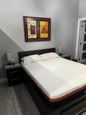 Uma cama bem arrumada com colcha branca, cabeceira de madeira e mesa de cabeceira com abajur.