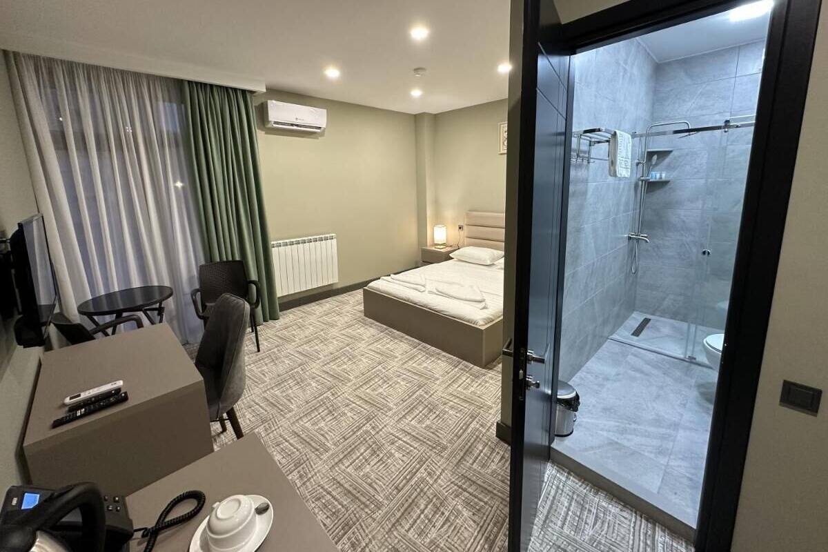 A hotel room with a bed, a desk with a chair, a TV, a telephone, and a shower area.