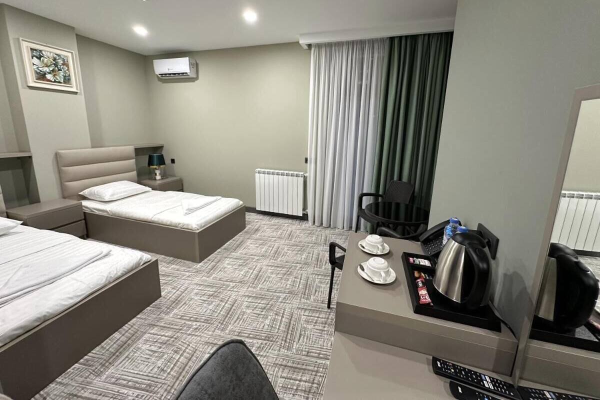A hotel room with two beds, a desk with a kettle and cups, a chair, a radiator, and a TV on the wall.